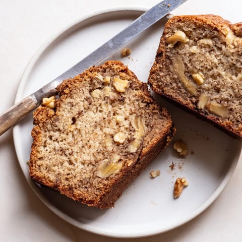 Freshly baked Banana Bread cooling on a wire rack, surrounded by ripe bananas and chopped walnuts for garnish.